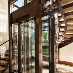 Curved glass-and-wood elevator enclosure beside a floating wood staircase, with aspen trees visible through the glass above.