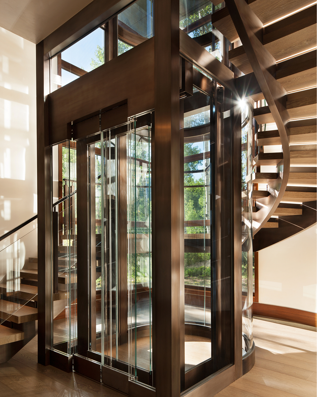 Curved glass-and-wood elevator enclosure beside a floating wood staircase, with aspen trees visible through the glass above.