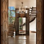View through open carved wood doors toward a glass elevator tower and curved floating wood staircase, lit by a cylindrical pendant chandelier, with aspen trees visible beyond the glass