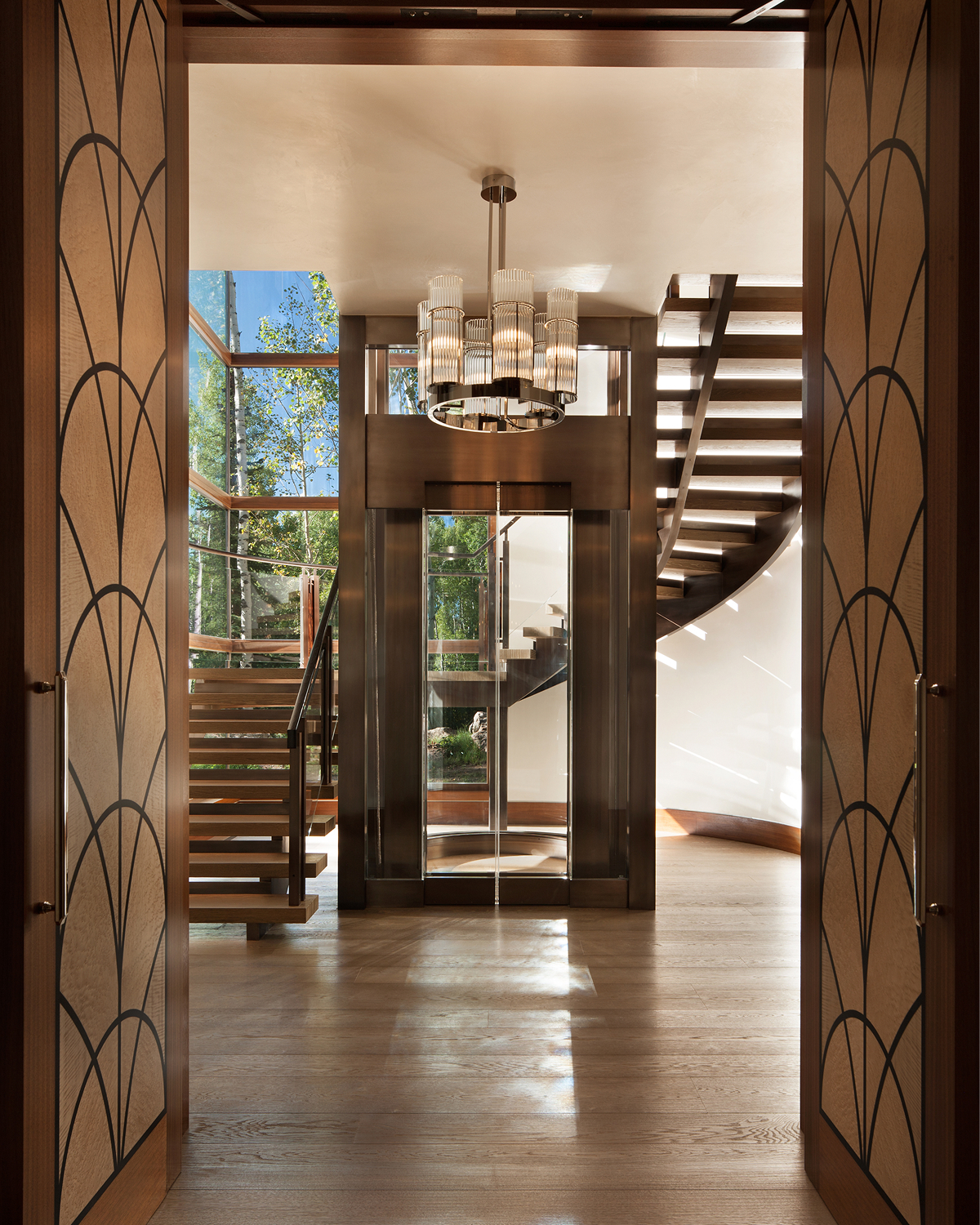 View through open carved wood doors toward a glass elevator tower and curved floating wood staircase, lit by a cylindrical pendant chandelier, with aspen trees visible beyond the glass