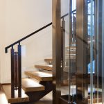 Floating wood staircase with open treads and a dark metal handrail, set against glass elevator panels