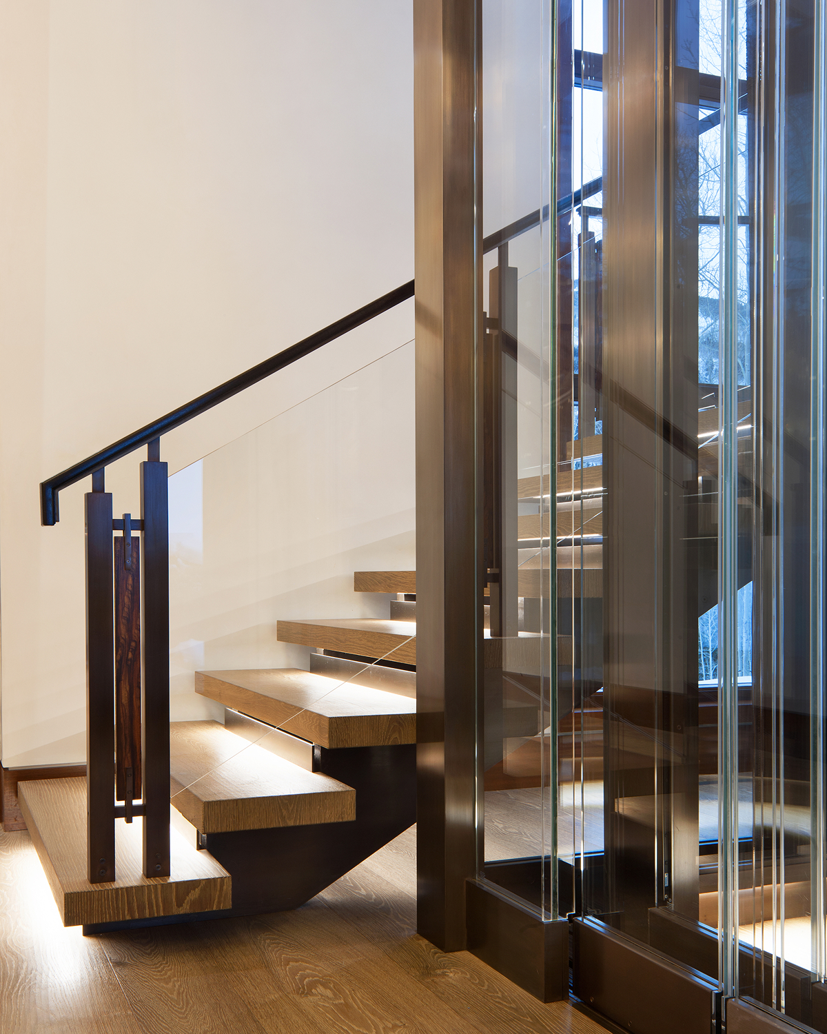 Floating wood staircase with open treads and a dark metal handrail, set against glass elevator panels