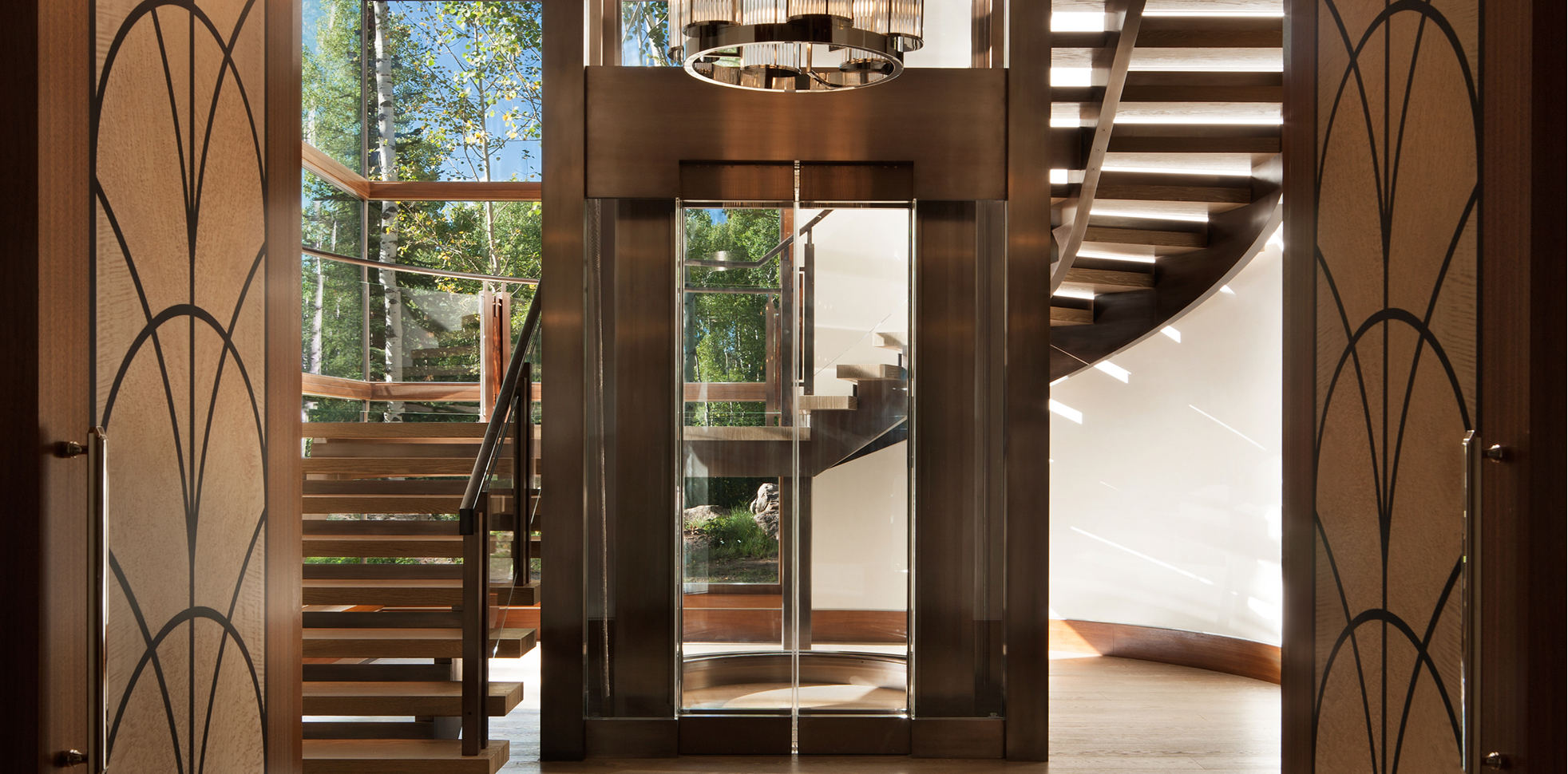 Wide view through carved wood doors of a glass elevator tower and curved floating wood staircase, lit by a cylindrical pendant chandelier, framed by birch trees outside
