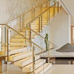 Floating staircase with illuminated onyx treads and a glass and brass railing, set against a bookmatched marble wall with a green stone sculpture and gravel art installation nearby