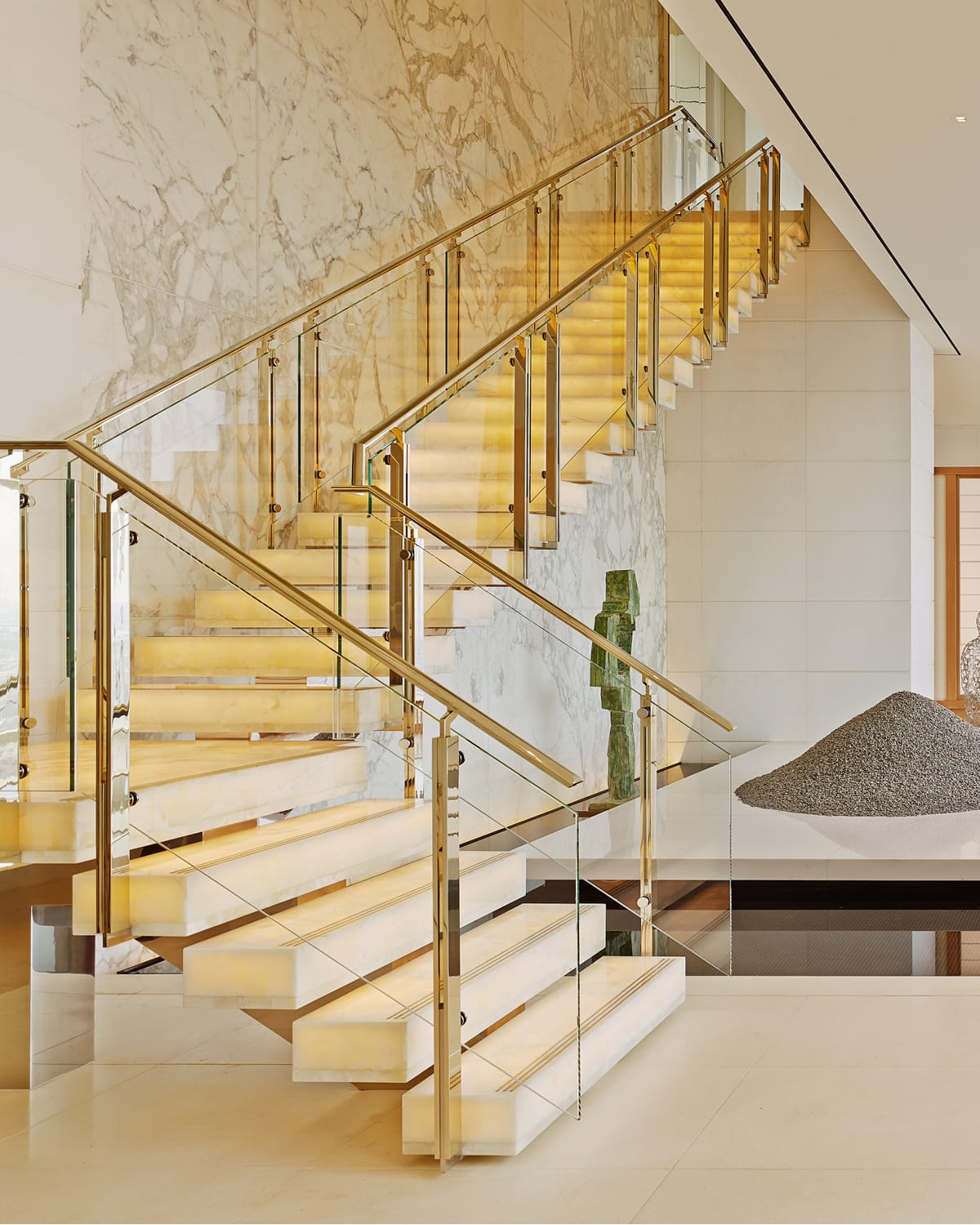 Floating staircase with illuminated onyx treads and a glass and brass railing, set against a bookmatched marble wall with a green stone sculpture and gravel art installation nearby