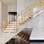 Floating staircase with illuminated onyx treads and a glass and brass railing, mirrored in a dark reflective floor beneath a bookmatched marble wall