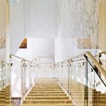 View looking down a staircase with illuminated onyx-clad treads and glass and brass railings, framed by floor-to-ceiling marble walls