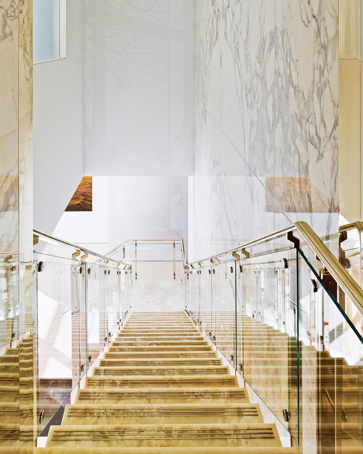 View looking down a staircase with illuminated onyx-clad treads and glass and brass railings, framed by floor-to-ceiling marble walls