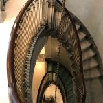 Looking down through an oval stairwell opening, framed by a dark wood handrail and brass-collared spindle railing, with grey marble treads curving around two visible flights and a chandelier chain dropping through the center