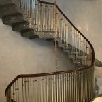 Curved staircase with a dark wood handrail and brass-collared spindle railing, descending along grey marble treads against a softly plastered wall