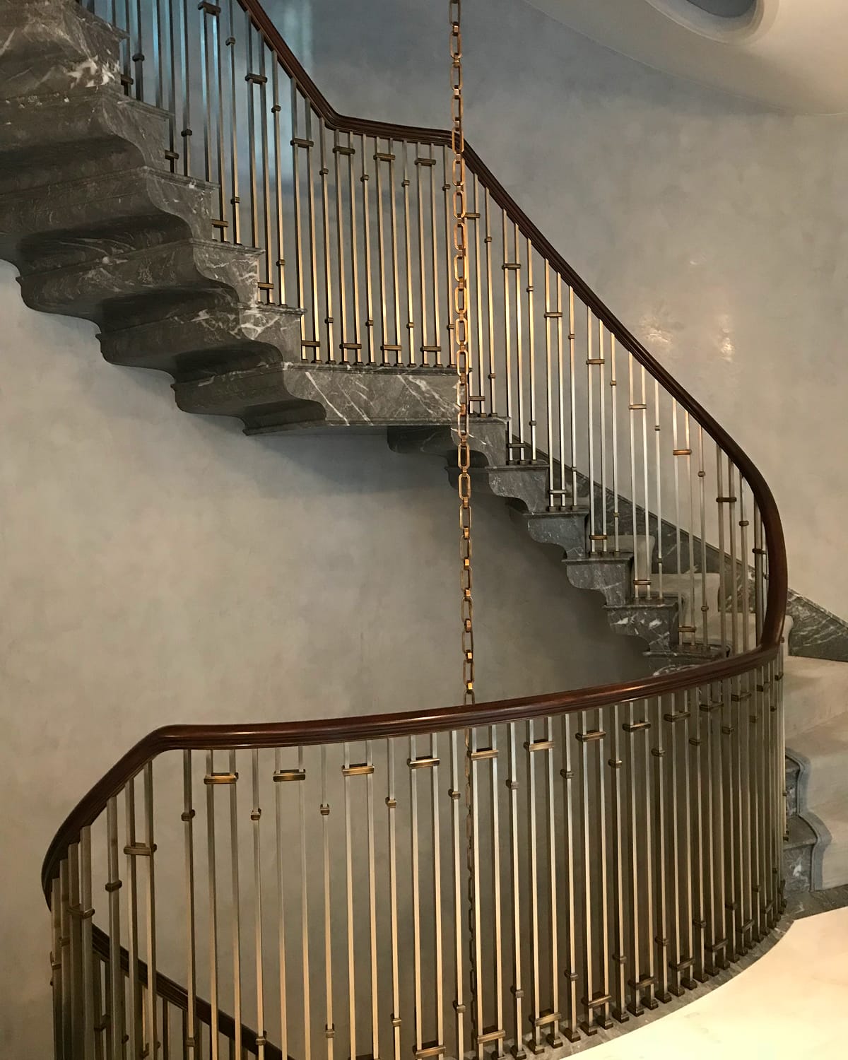 Curved staircase with a dark wood handrail and brass-collared spindle railing, descending along grey marble treads against a softly plastered wall