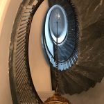 Looking straight up through an oval stairwell lined in grey marble and spindle railing, toward a domed skylight, with a gilded chandelier in the foreground