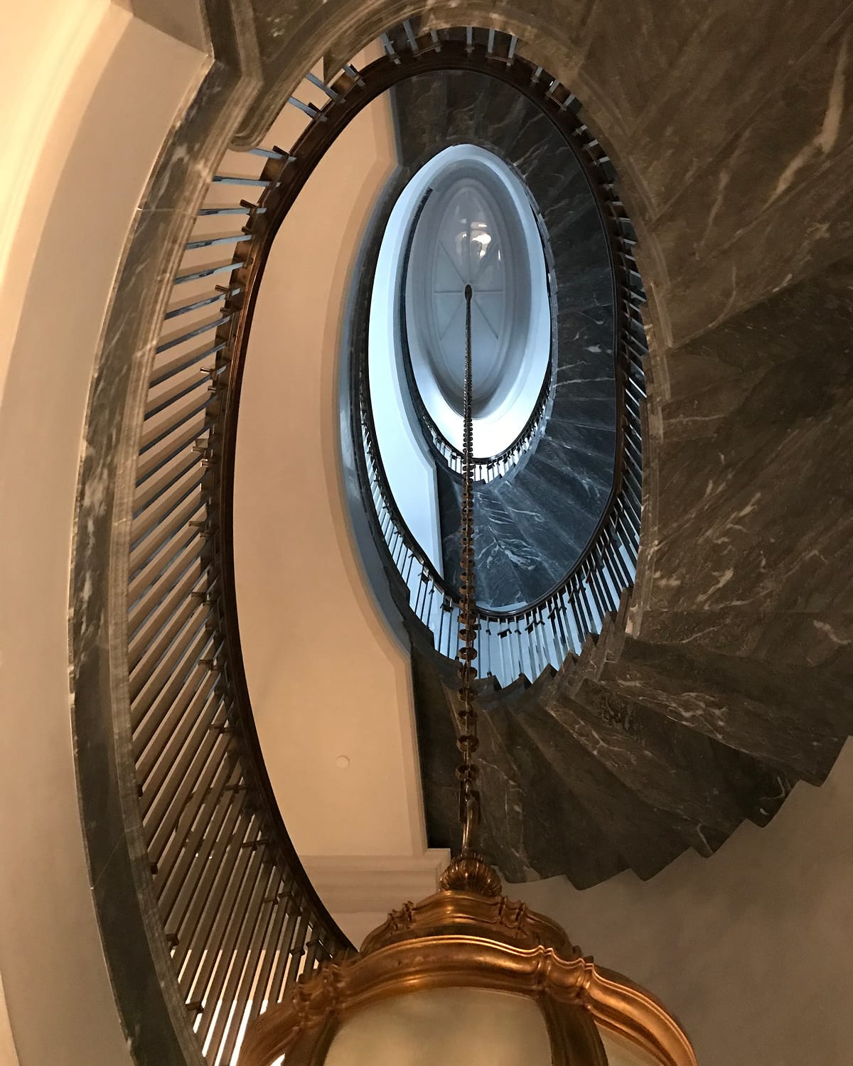 Looking straight up through an oval stairwell lined in grey marble and spindle railing, toward a domed skylight, with a gilded chandelier in the foreground