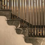 Close detail of scalloped grey marble stair treads and brass-collared spindle railing bases, lit warmly against a softly plastered wall