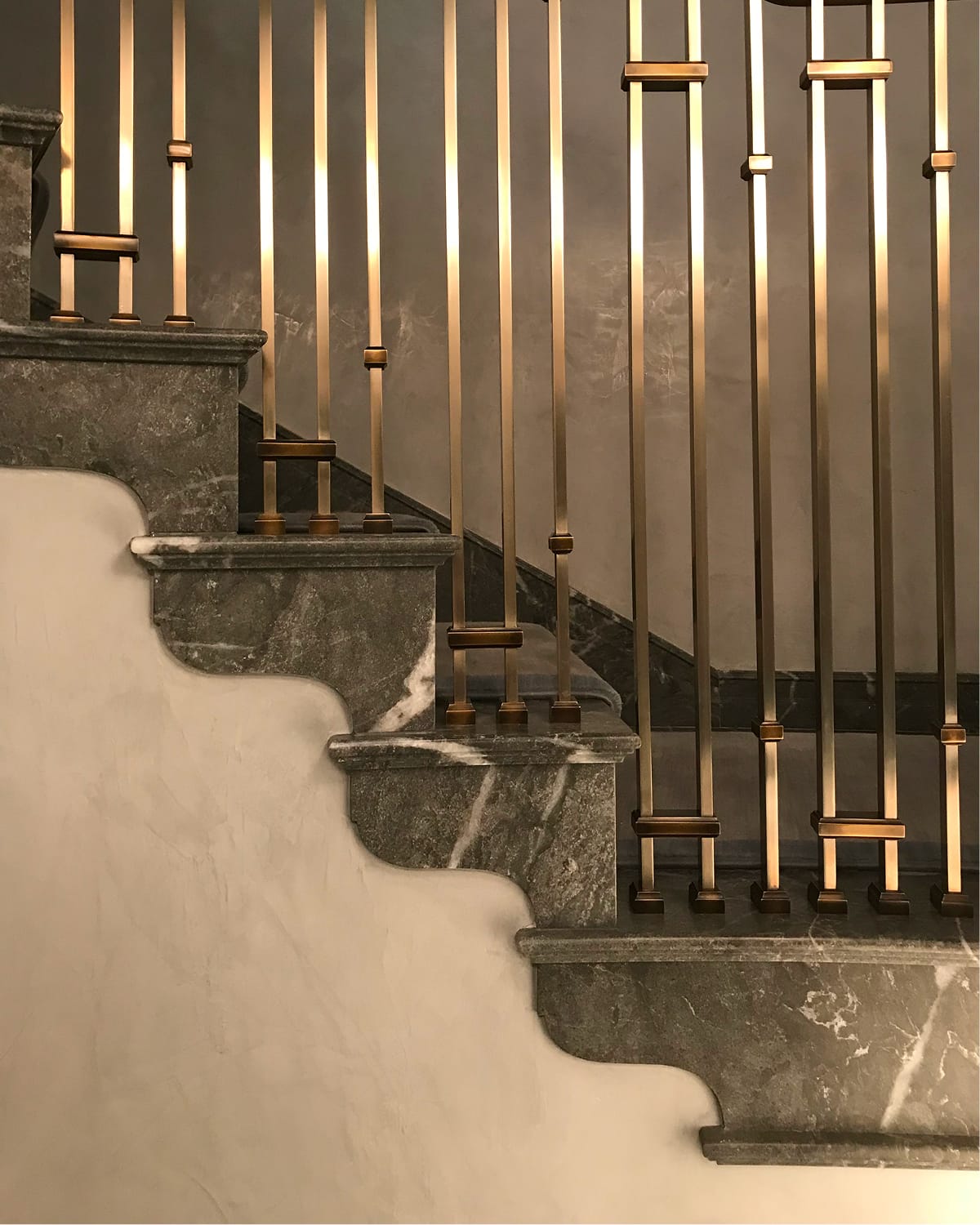 Close detail of scalloped grey marble stair treads and brass-collared spindle railing bases, lit warmly against a softly plastered wall