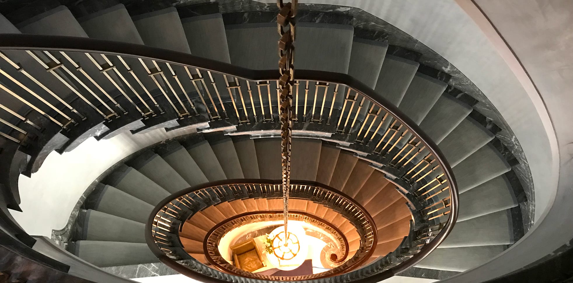 Overhead view down an oval staircase, its grey marble treads and brass-collared spindle railing spiraling around a pendant chandelier glowing at the base