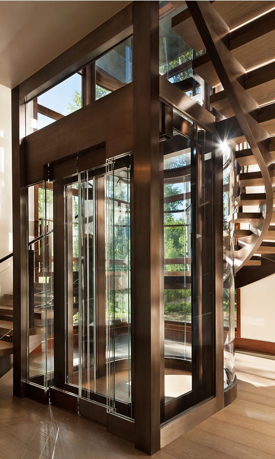 Glass and bronze-framed elevator shaft integrated beside a curved staircase with wood treads