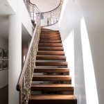 Floating staircase with dark wood treads and a metal railing cast in an organic, cell-like lattice pattern, curving up two stories beside a sunlit white stairwell