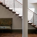 Floating staircase with dark wood treads and a metal railing cast in an organic, cell-like lattice pattern, seen past a round white column and an antique upholstered bench