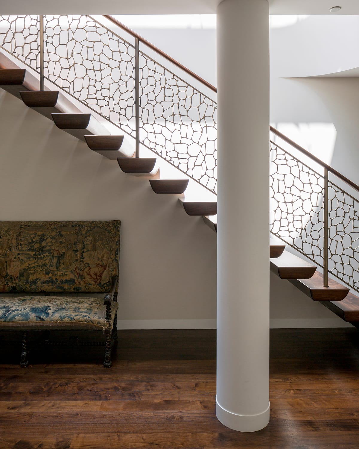 Floating staircase with dark wood treads and a metal railing cast in an organic, cell-like lattice pattern, seen past a round white column and an antique upholstered bench