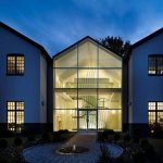 Glass-gabled entrance pavilion lit at night, revealing a sculptural crisscrossed metal installation surrounding a floating staircase inside, framed by a brick courtyard path and fountain