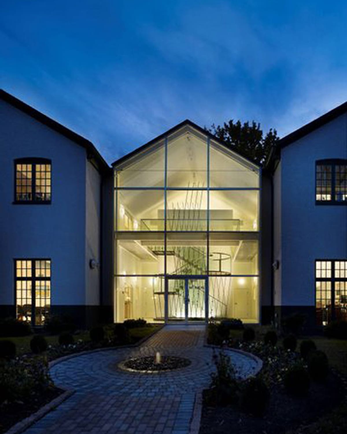 Glass-gabled entrance pavilion lit at night, revealing a sculptural crisscrossed metal installation surrounding a floating staircase inside, framed by a brick courtyard path and fountain