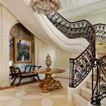 Curved staircase with scrolled iron and gilt-bronze railing beneath a crystal chandelier, in a gilded entry hall with patterned wallpaper, a gold-leafed pedestal table, and a mirrored alcove with a framed painting.