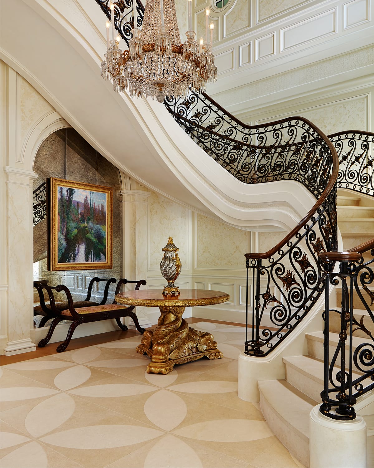 Curved staircase with scrolled iron and gilt-bronze railing beneath a crystal chandelier, in a gilded entry hall with patterned wallpaper, a gold-leafed pedestal table, and a mirrored alcove with a framed painting.