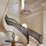 Curved staircase with scrolled iron and gilt-bronze railing descending into a two-story entry hall, lit by a tall crystal chandelier, with a gold-leafed pedestal table at the base