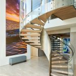 Spiral staircase with light wood treads and a glass and brushed-metal railing, curving past a large sunset seascape photograph and a bench in a bright foyer