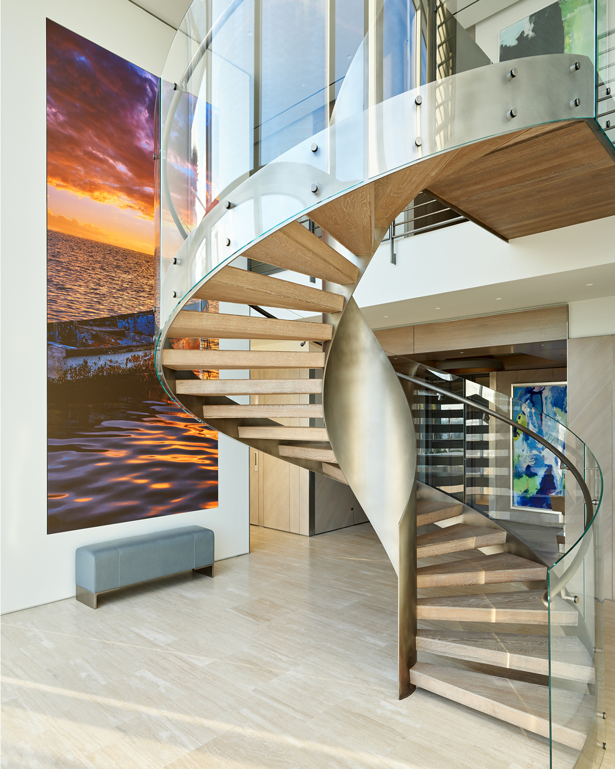 Spiral staircase with light wood treads and a glass and brushed-metal railing, curving past a large sunset seascape photograph and a bench in a bright foyer