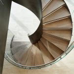 Overhead view of a spiral staircase, showing light wood treads radiating around a curved brushed-metal center spine
