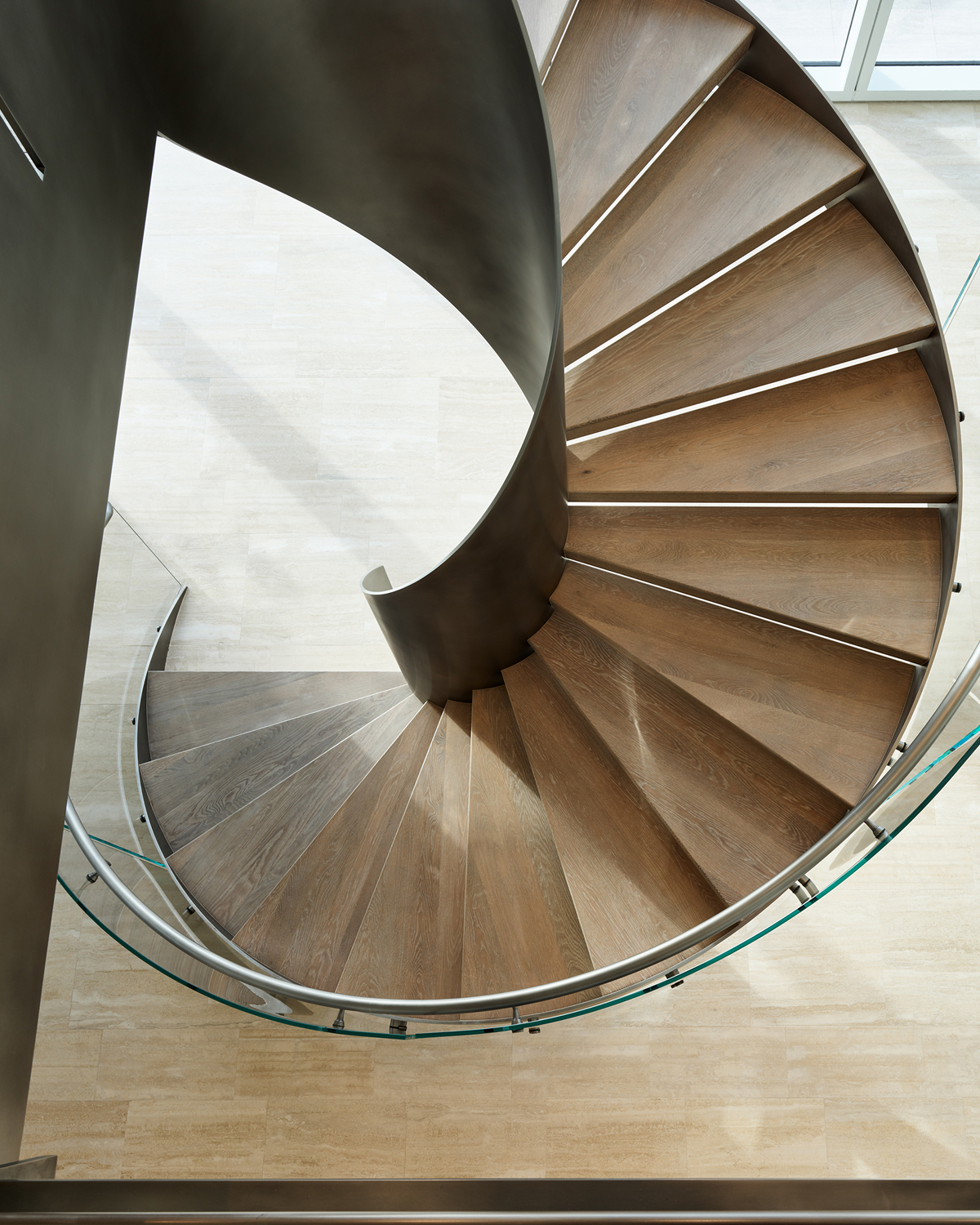 Overhead view of a spiral staircase, showing light wood treads radiating around a curved brushed-metal center spine