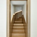 View down a narrow wood-paneled stairwell toward a curved staircase with light wood treads and an illuminated handrail channel