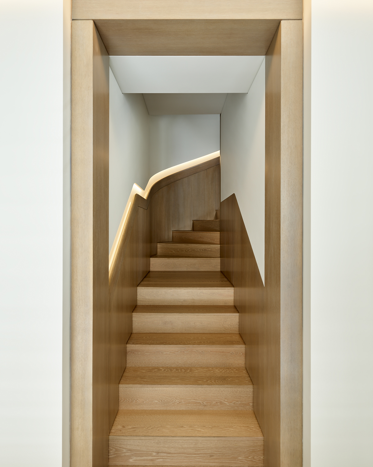 View down a narrow wood-paneled stairwell toward a curved staircase with light wood treads and an illuminated handrail channel