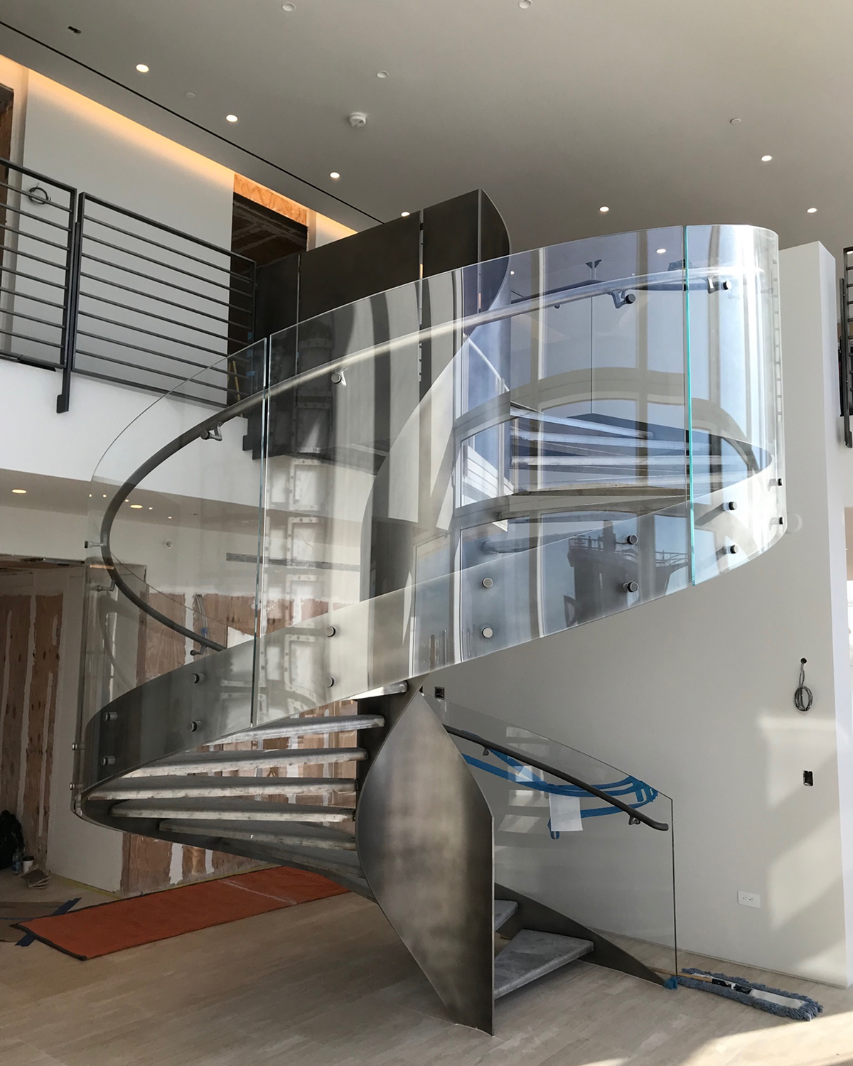 In-progress installation of a spiral staircase, showing the brushed-metal structure and glass railing partially complete, with construction materials and floor protection visible