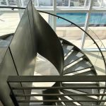 Spiral staircase with wood treads and a glass and brushed-metal railing, beside floor-to-ceiling windows