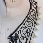 Upward view along a spiral staircase's scrolled iron railing, curving toward an oval domed ceiling with a sunburst chandelier and marble treads.