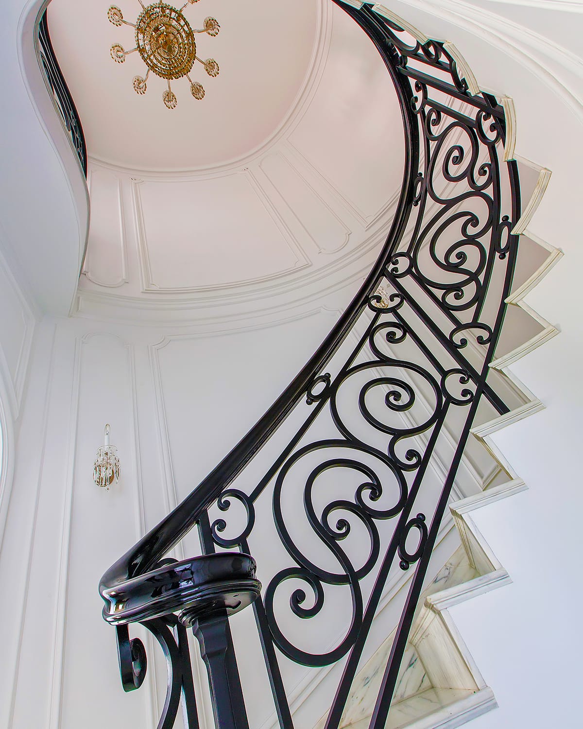 Upward view along a spiral staircase's scrolled iron railing, curving toward an oval domed ceiling with a sunburst chandelier and marble treads.