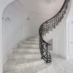 Spiral staircase with white marble treads and a scrolled iron railing, curving up from a marble landing in an all-white paneled rotunda