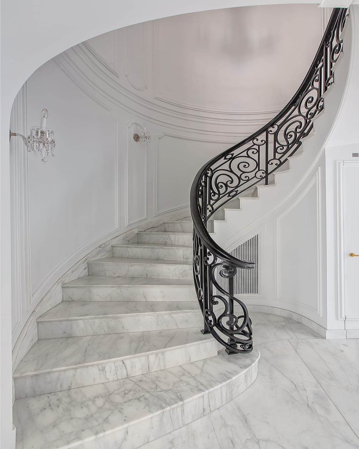 Spiral staircase with white marble treads and a scrolled iron railing, curving up from a marble landing in an all-white paneled rotunda