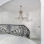 Top landing of a spiral staircase, with a scrolled iron railing framing the opening beneath a crystal chandelier and marble flooring