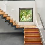 Floating staircase with wood treads edged in a rust-orange accent and a glass railing, next to a square window framing green foliage