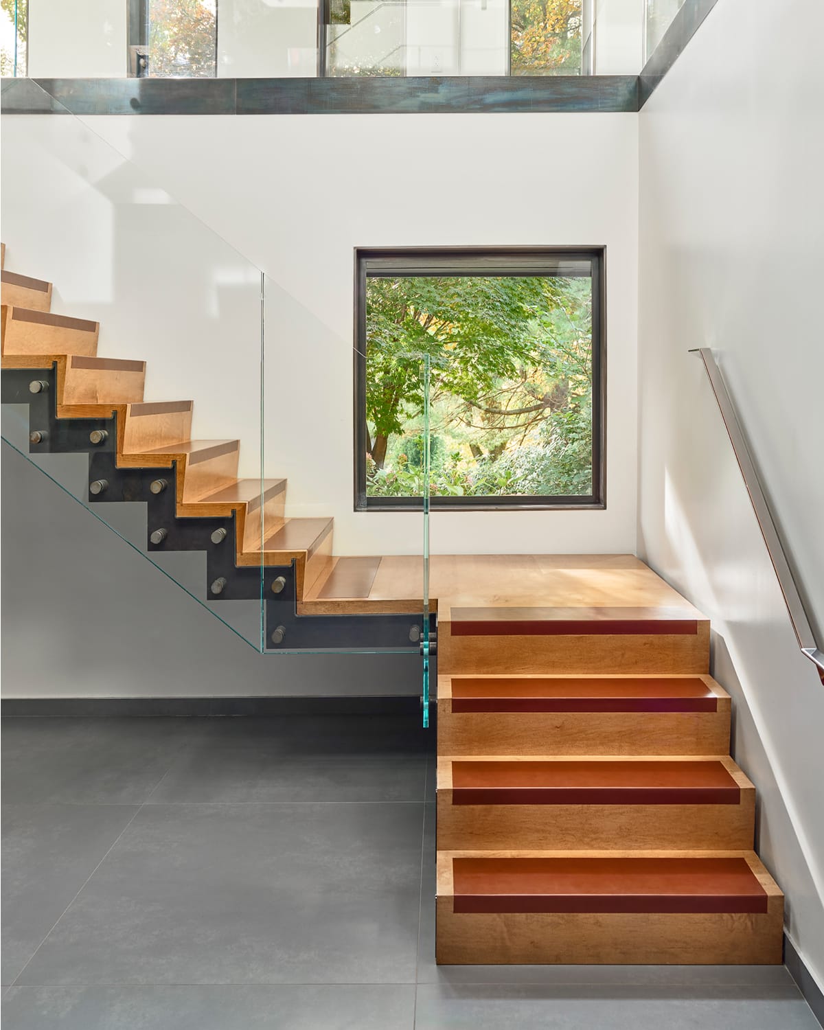 Floating staircase with wood treads edged in a rust-orange accent and a glass railing, next to a square window framing green foliage