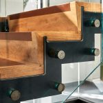 Close detail of wood stair treads with rust-orange accent edges, joined to an exposed steel stringer and glass railing panel with visible mounting bolts