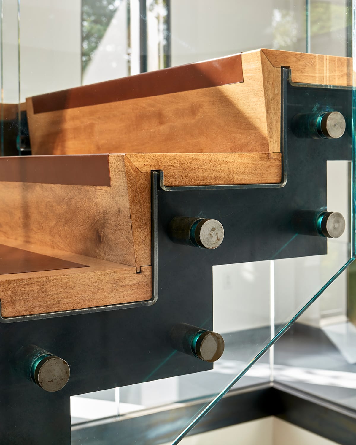 Close detail of wood stair treads with rust-orange accent edges, joined to an exposed steel stringer and glass railing panel with visible mounting bolts