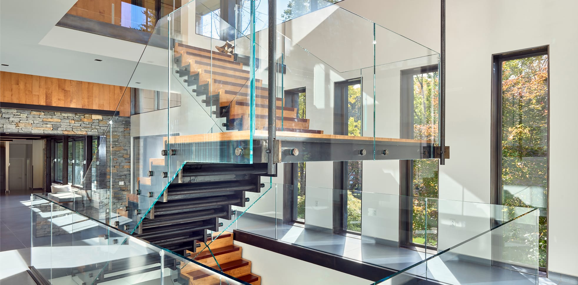 Floating staircase with wood treads, dark steel supports, and a glass railing, spanning an open two-story space with a stone fireplace wall and tall windows beyond.