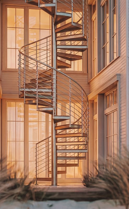 Outdoor spiral staircase on a coastal home, with multi-line railing and dune grass in the foreground at sunset