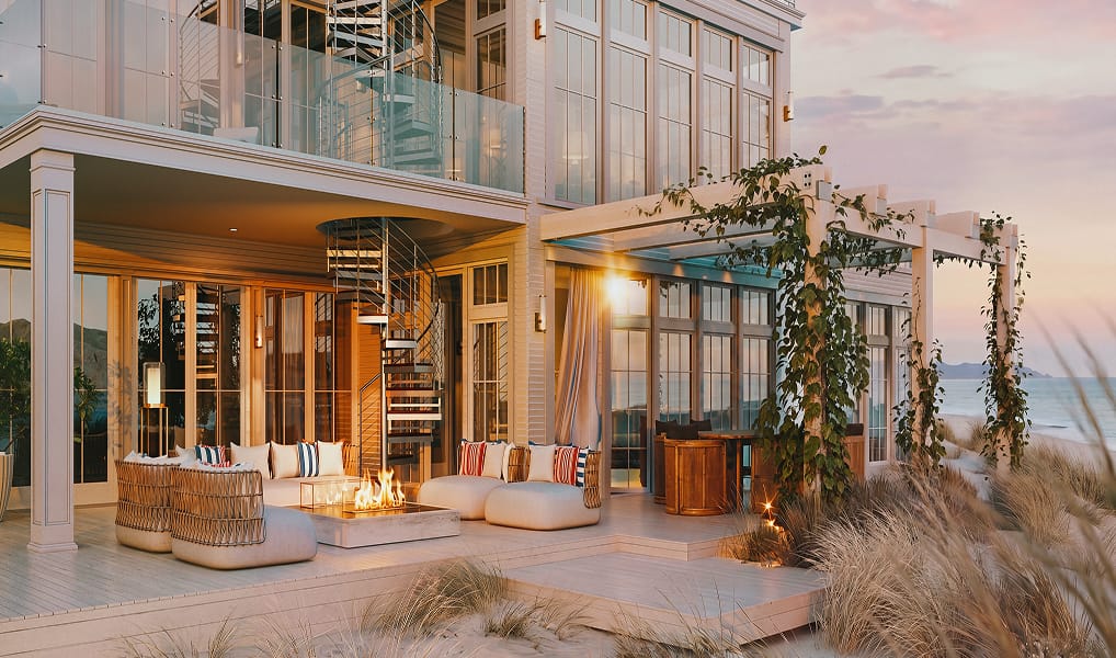 Coastal home exterior with a metal spiral staircase on the balcony, outdoor lounge seating, and a fire pit at sunset