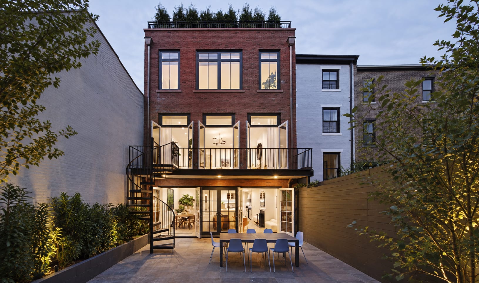 Black exterior metal spiral staircase on a brick rowhouse, connecting a balcony to a lit patio with dining table at dusk